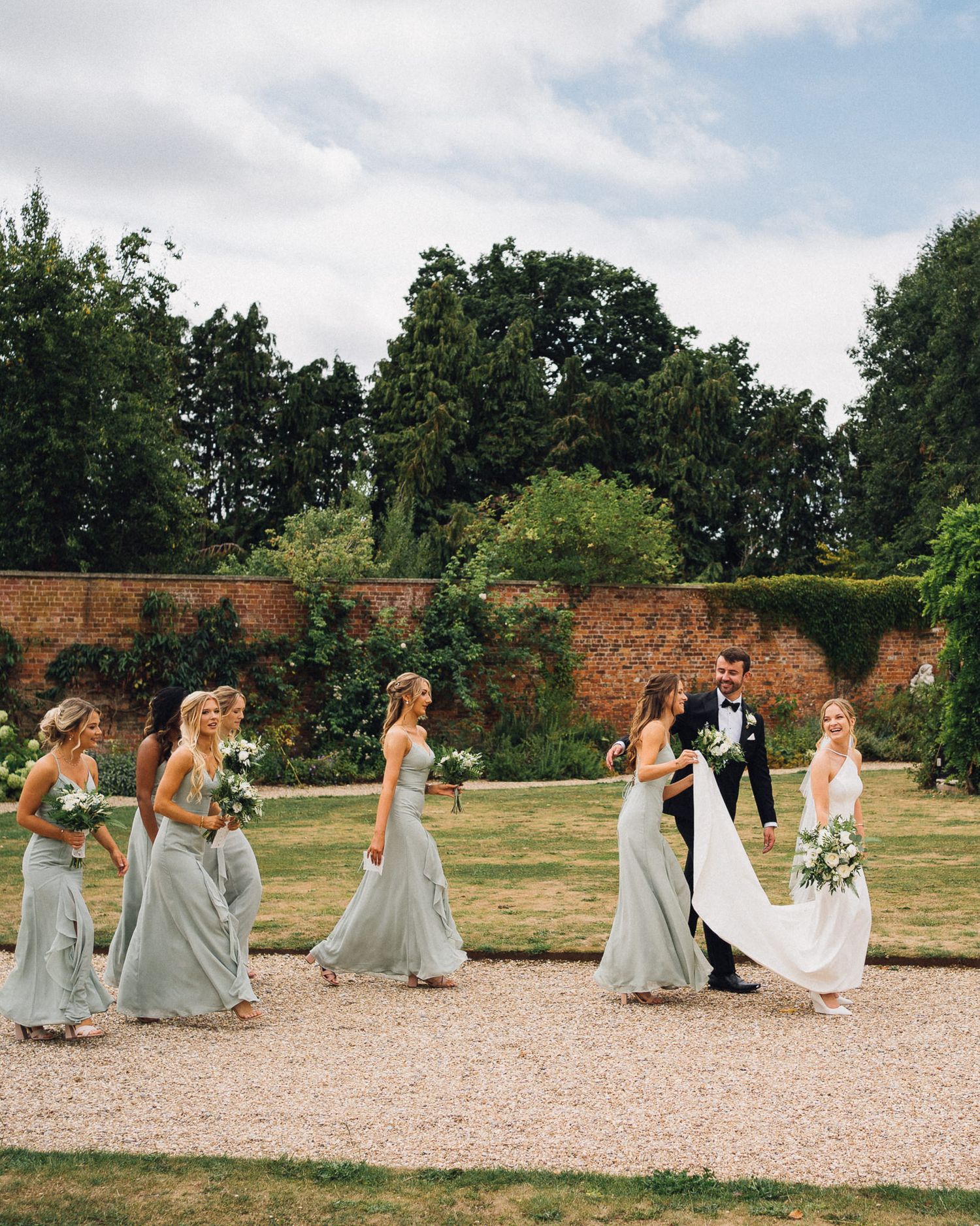 Bridal party in gray dresses walking across gravel path near historic brick wall.