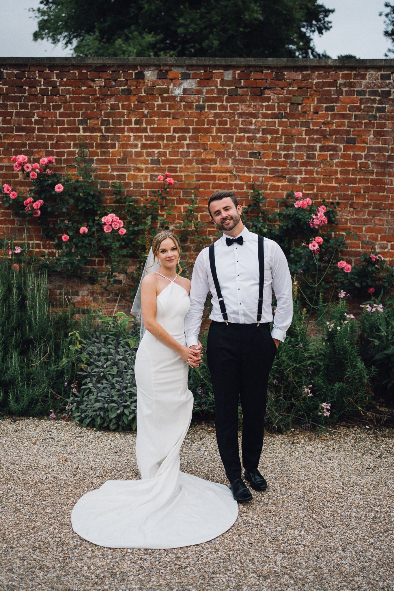 A couple poses for wedding photos in front of a brick wall adorned with pink roses.