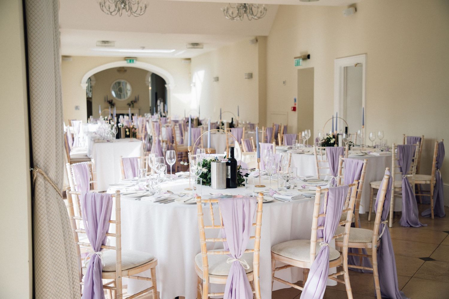 Wedding reception hall setup with white round tables and chairs adorned with lavender sashes.
