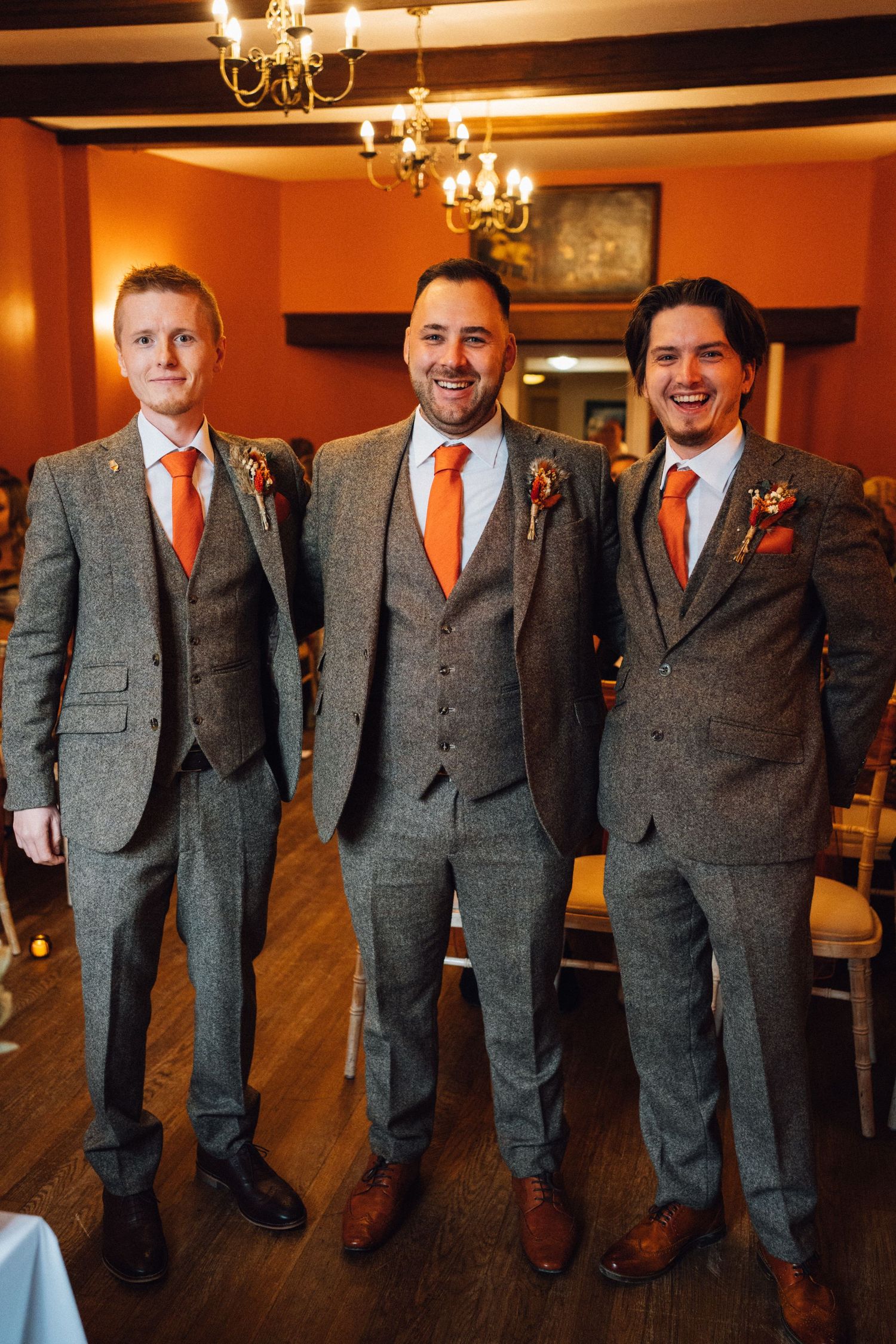 Groomsmen in matching gray tweed suits and orange ties stand together in an elegant indoor venue.