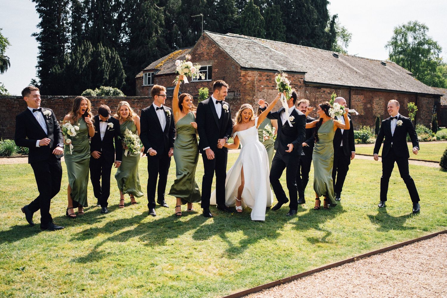 Wedding group poses playfully on grassy lawn in front of rustic brick buildings.