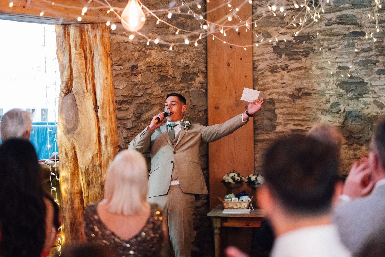 A person in a light suit enthusiastically addresses wedding guests during a reception in a rustic venue with string lights.