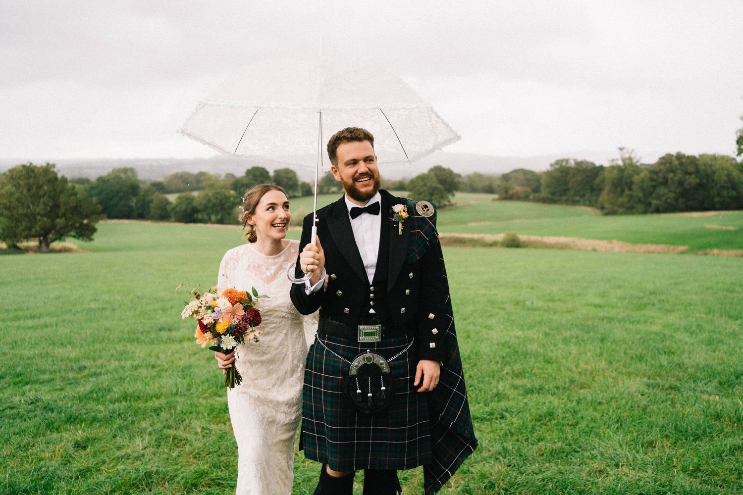 Smiling bride in a white gown holding a bouquet walks with a groom in a kilt and tartan sash under a clear umbrella across a green field.