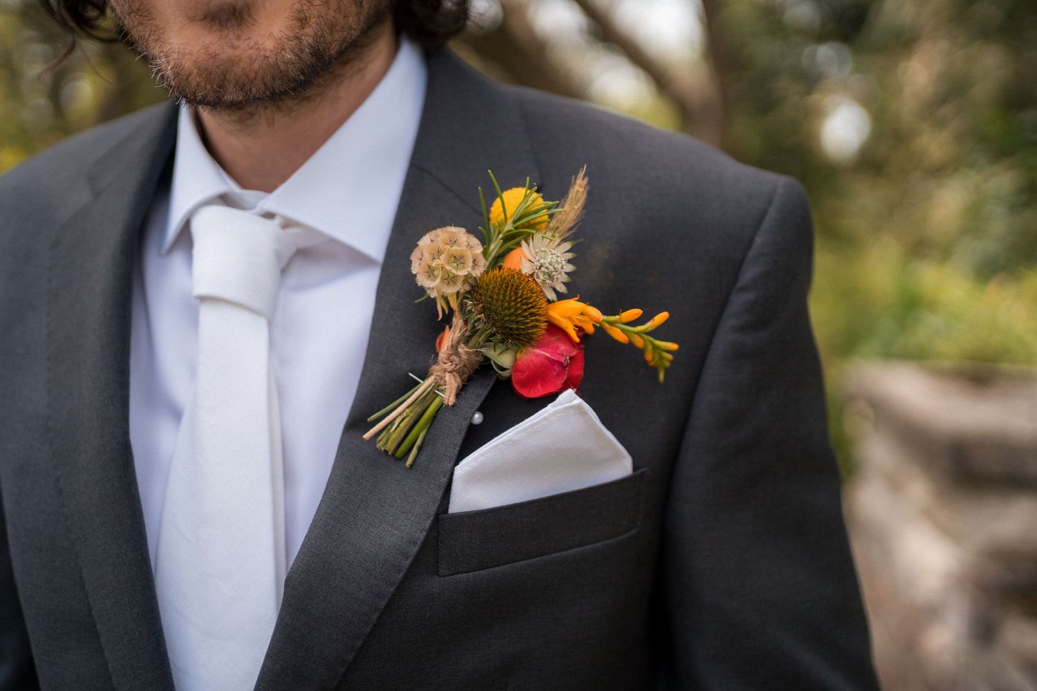 Close-up of a groom's charcoal suit boutonniere featuring vibrant wildflowers, a white pocket square, and white tie.