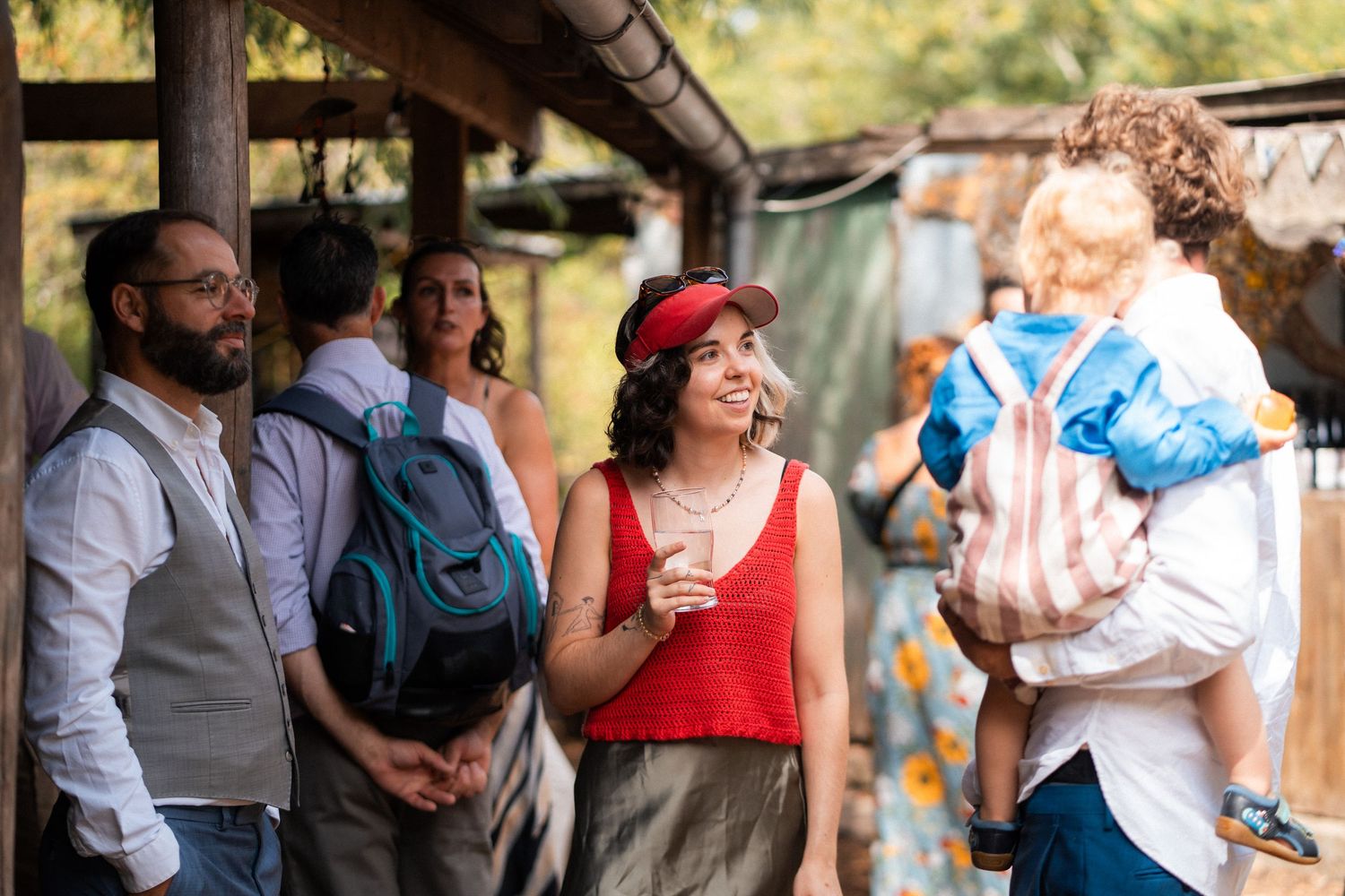 Group of diverse friends socializing outdoors at a rustic venue, woman in red top and cap smiling.