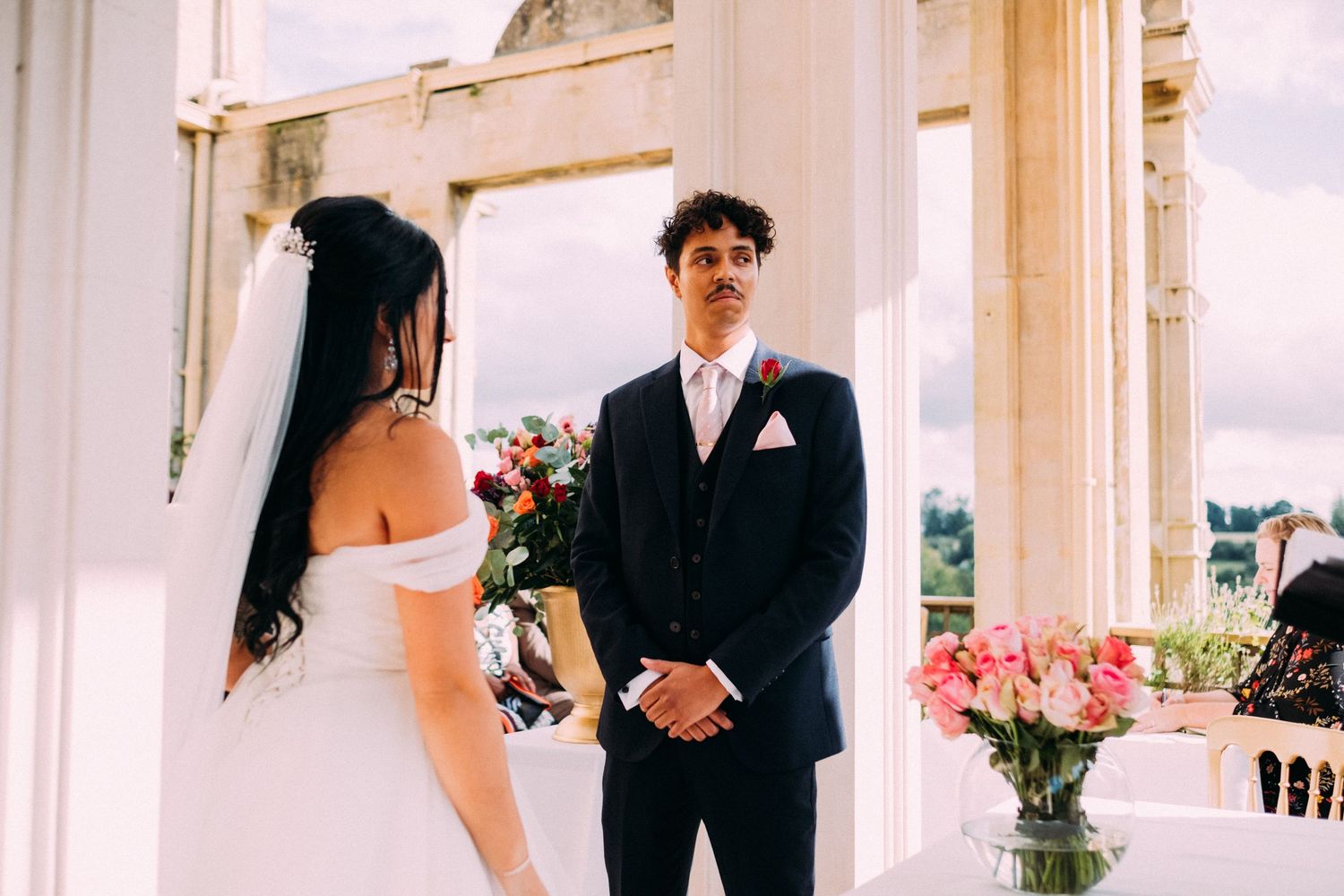 A bride and groom share a moment during their outdoor wedding ceremony beside pink floral arrangements.