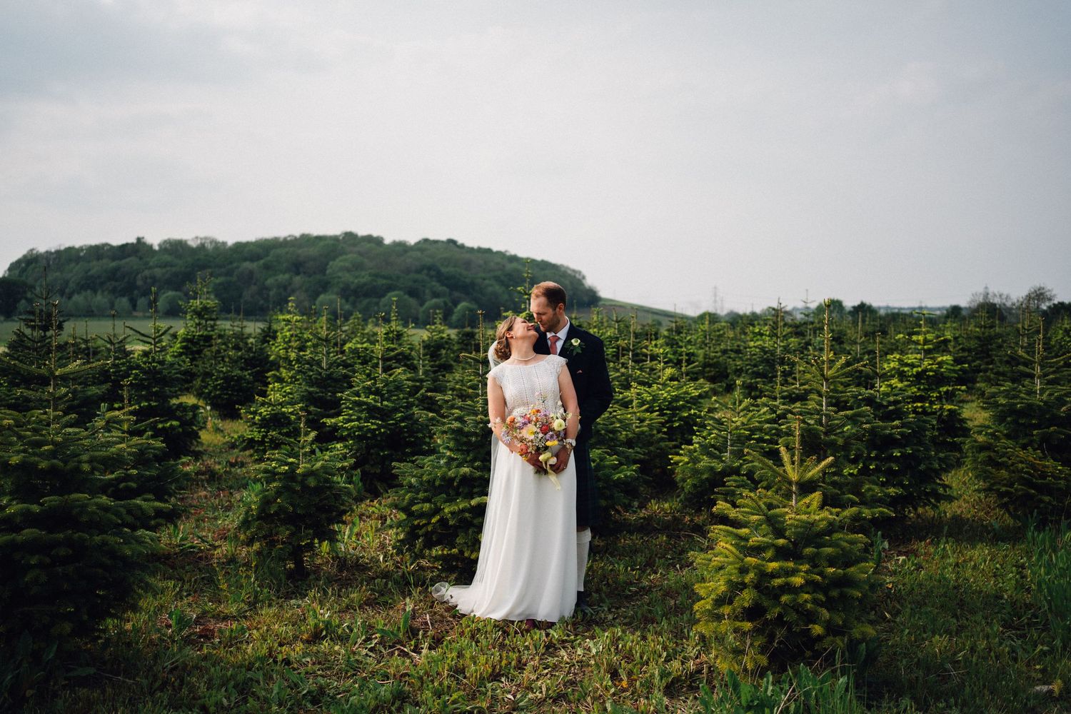 Wedding couple poses against scenic mountain landscape with evergreen trees and cloudy sky.