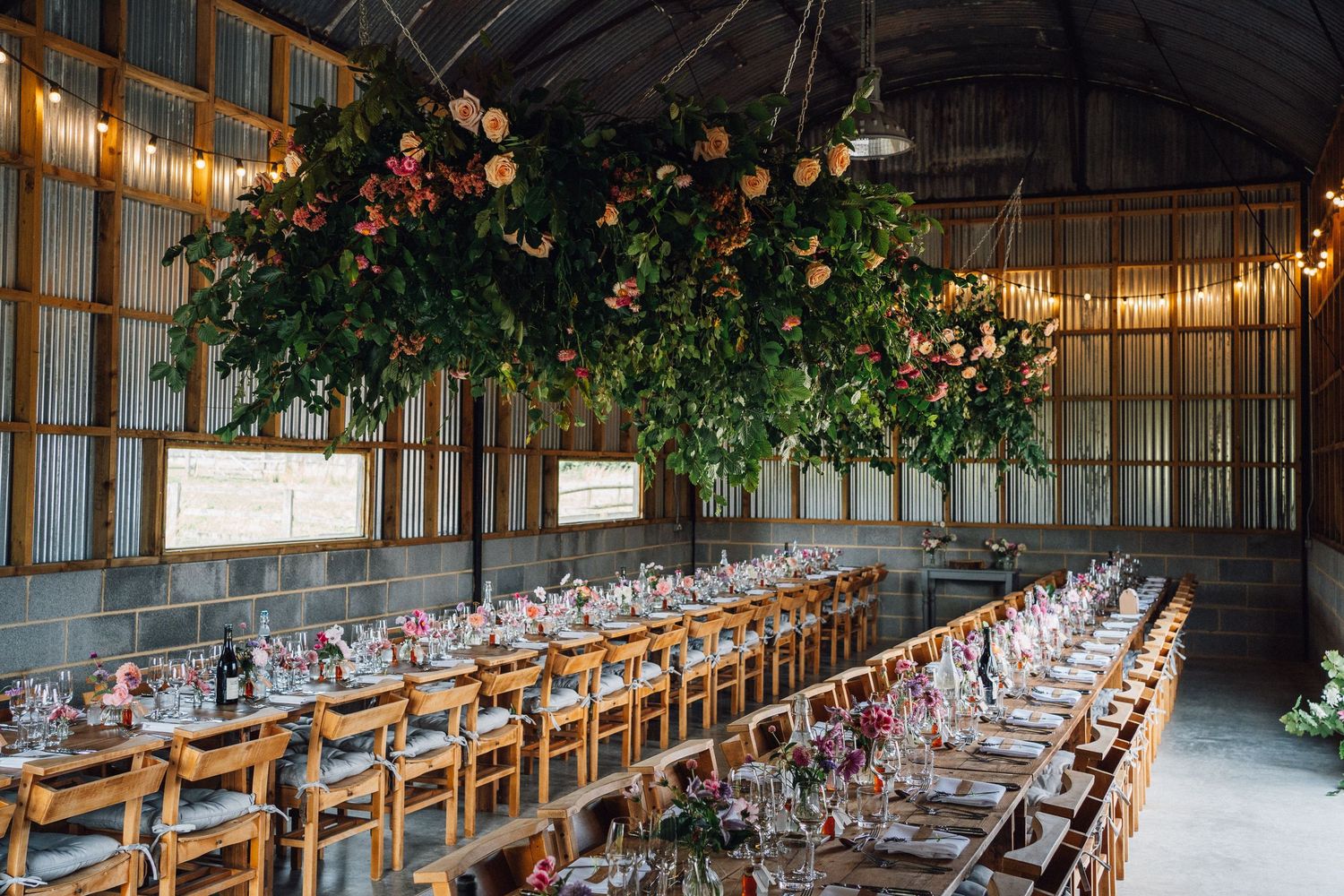 Elegant table settings in a barn venue with suspended greenery and warm lighting create a romantic atmosphere.