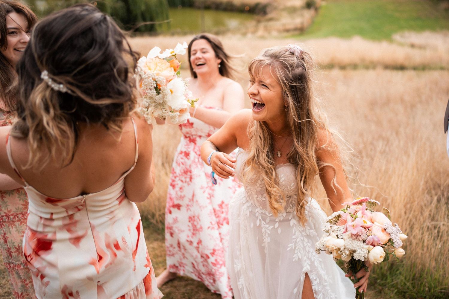 Joyful bride laughing with bridesmaids in floral dresses holding bouquets in a golden field.