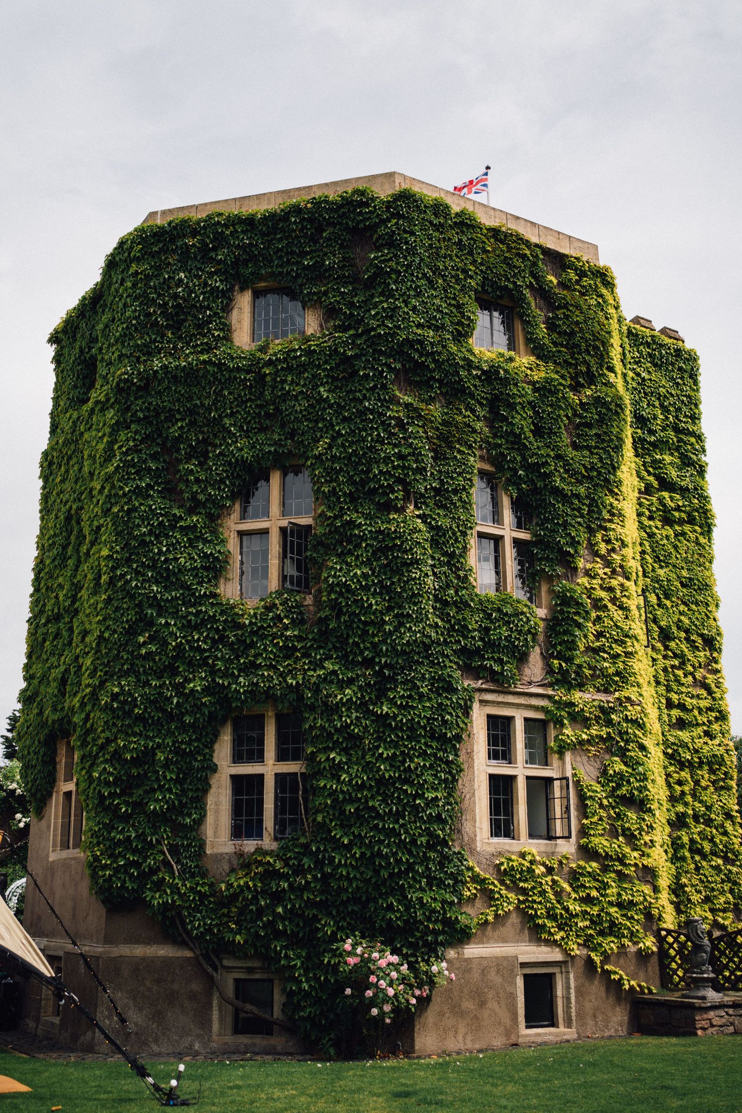 Historic stone building covered in lush green ivy with a Union Jack flag on top, set against an overcast sky.