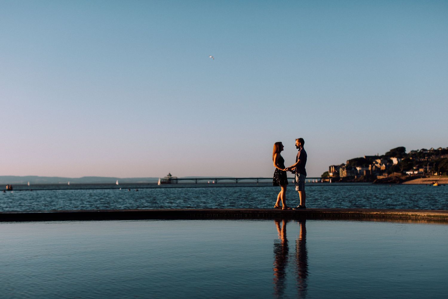 Two silhouettes stand together on a beach at sunset with calm ocean waters in the background.