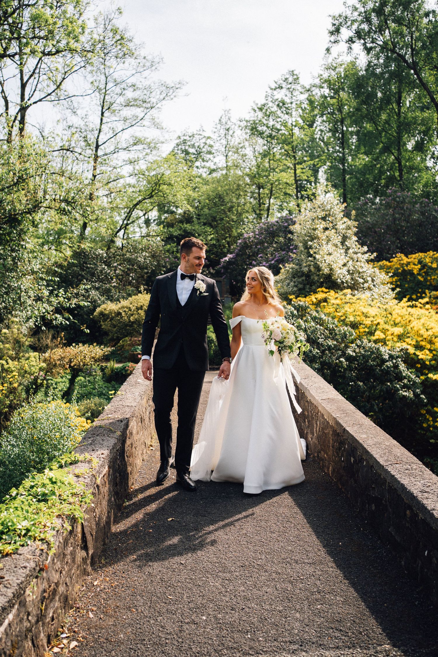 A couple walks along a stone bridge path surrounded by lush green trees and yellow flowers on their wedding day.