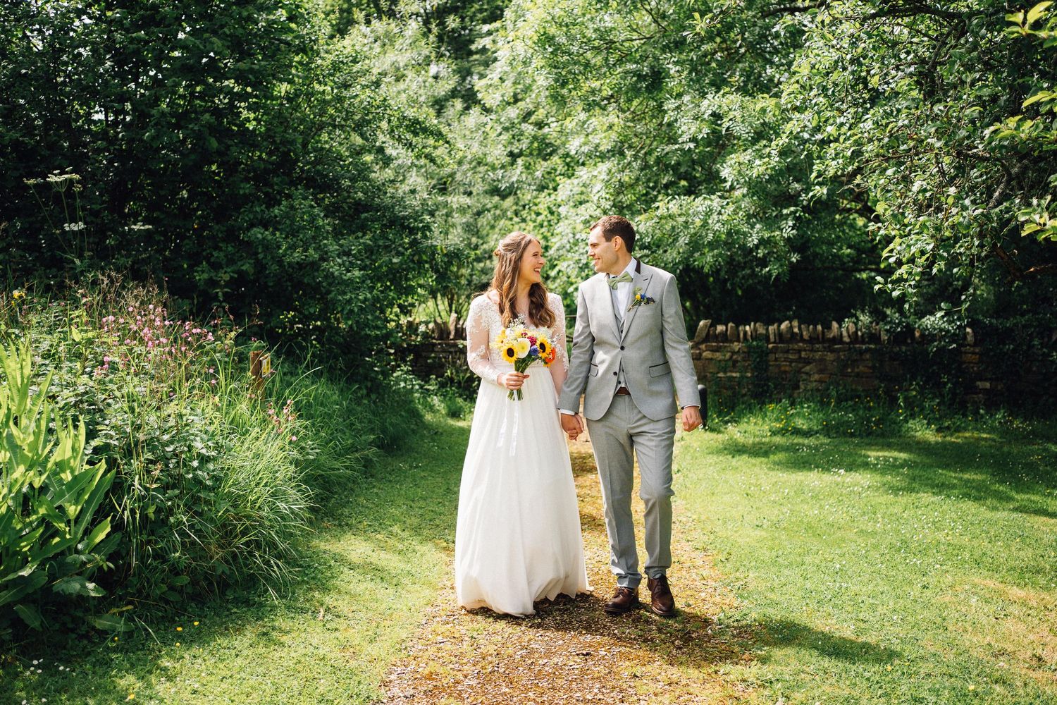 Wedding couple walks hand in hand through a lush garden path surrounded by green foliage on their special day.