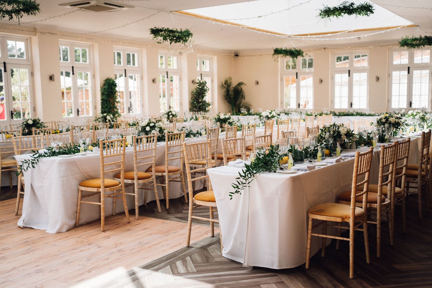 Elegant wedding reception venue with natural light, white tablecloths, wooden chairs and greenery decorations throughout.