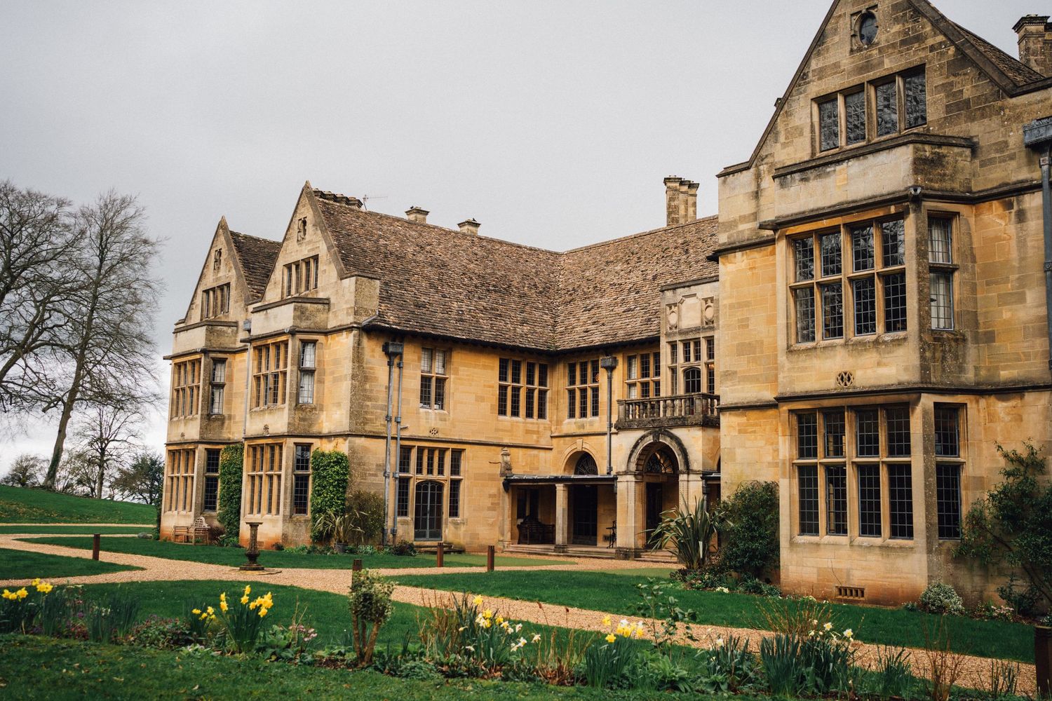 Grand Elizabethan-style manor house with ornate windows and chimneys surrounded by a manicured lawn and daffodils.