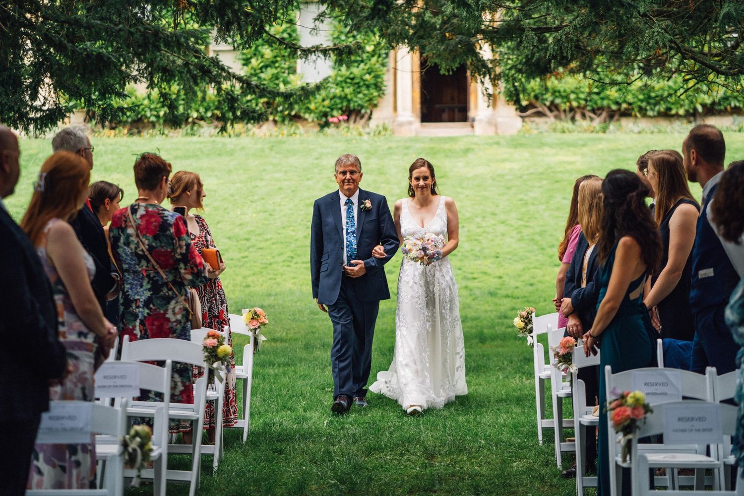 Bride walks down outdoor aisle with escort at garden wedding ceremony with white chairs.