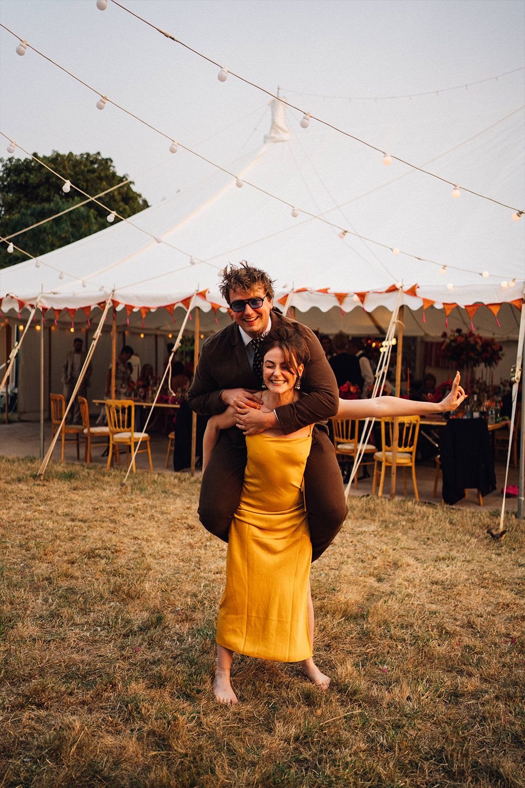 A couple dances together under string lights at an outdoor wedding reception with a tent in the background.