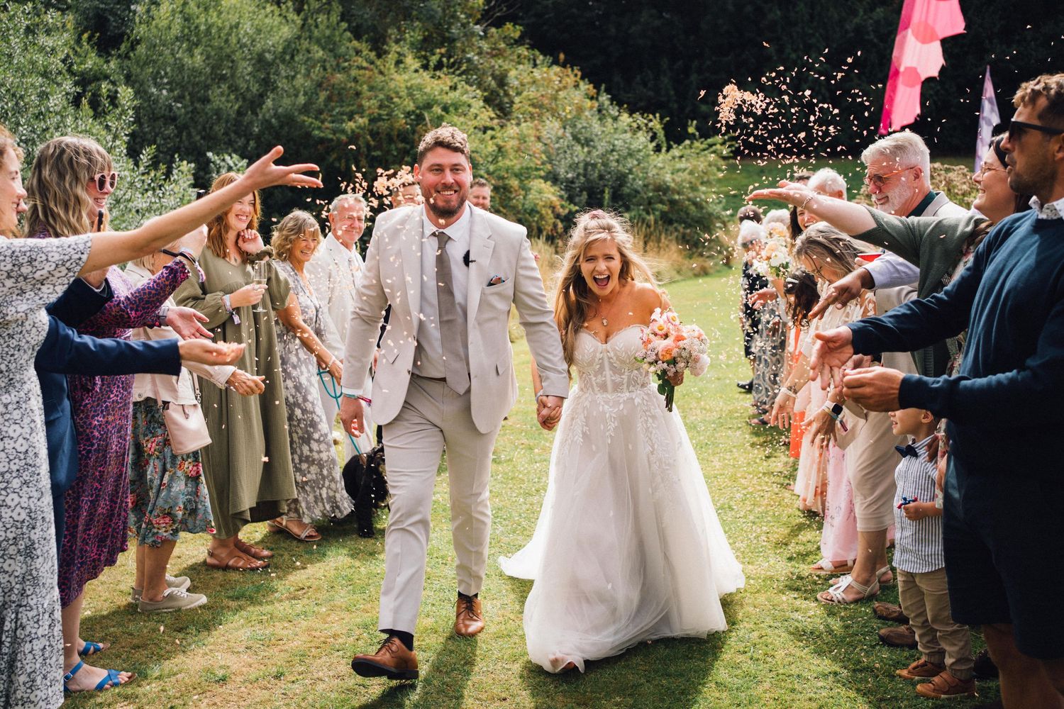 Wedding guests throw confetti and celebrate on a grassy lawn during an outdoor summer celebration.
