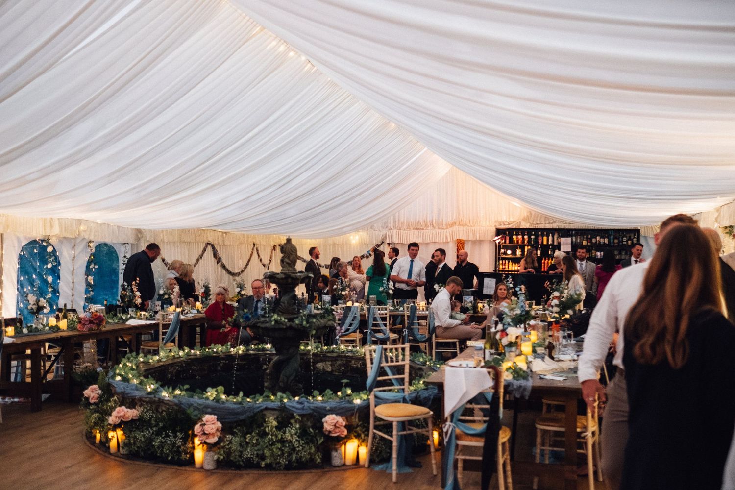 Elegant wedding reception setup in a white tent with hanging lights and floral centerpieces on dining tables.