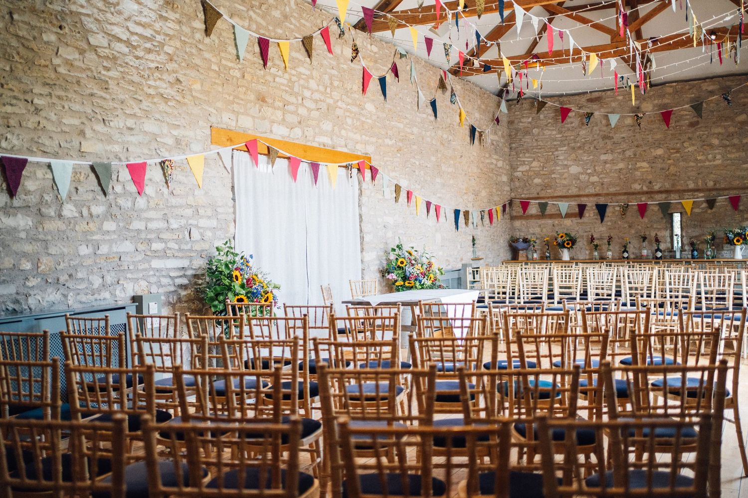 Rustic barn wedding venue with colorful bunting, wooden chairs in rows, stone walls, and floral arrangements.