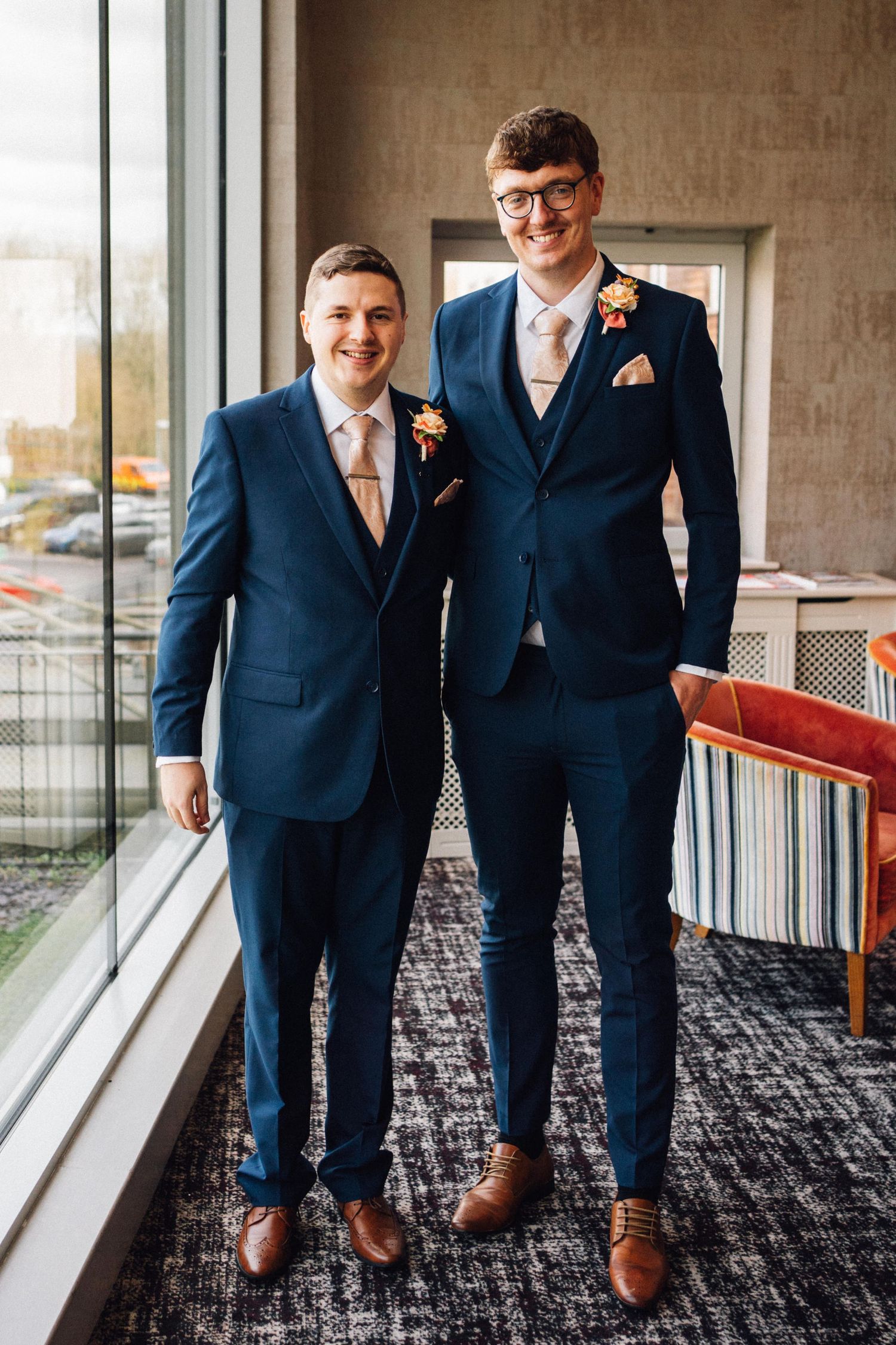 Close-up shots of navy blue suits and brown dress shoes on a patterned carpet by large windows.