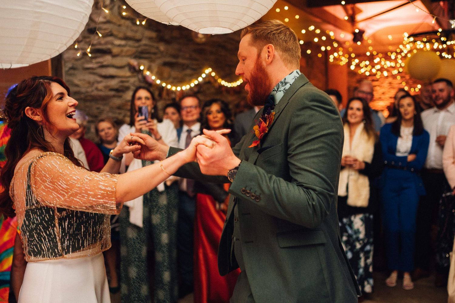 Wedding reception dance floor filled with guests celebrating under glowing string lights and paper lanterns.