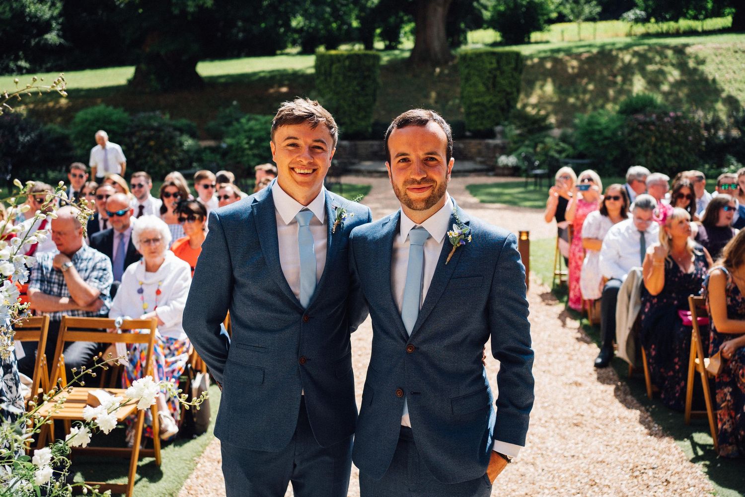 Wedding party members in blue suits and light blue dresses pose outdoors in a garden setting.