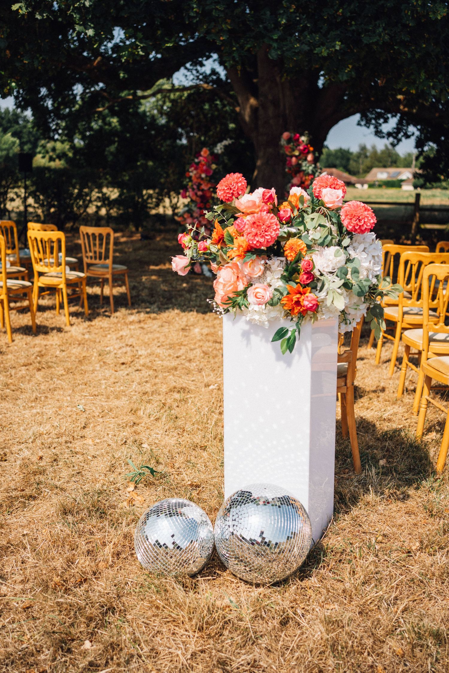 Outdoor wedding ceremony setup with coral floral arrangement on white pedestal and disco balls against rows of wooden chairs.