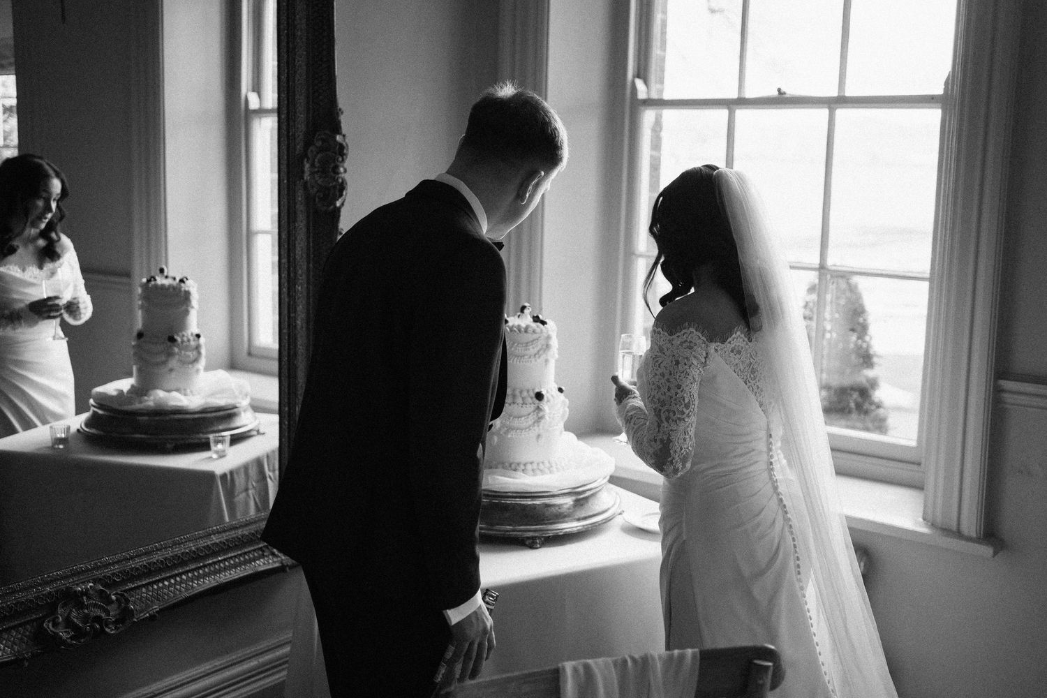 Black and white photo of bride and groom cutting their wedding cake by a bright window.