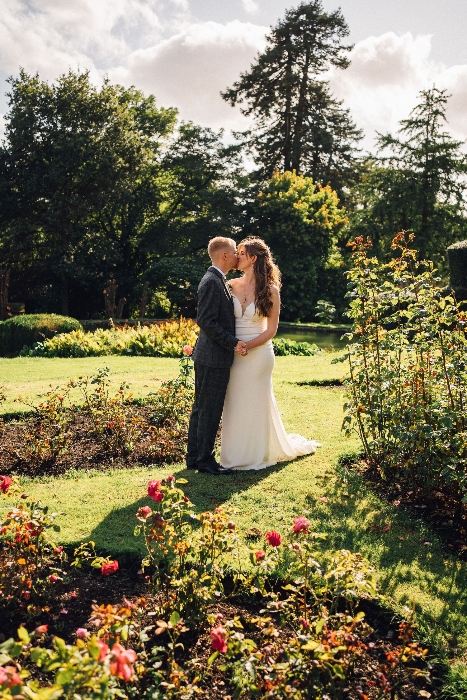Newlyweds pose in garden with blooming roses and golden sunset light.