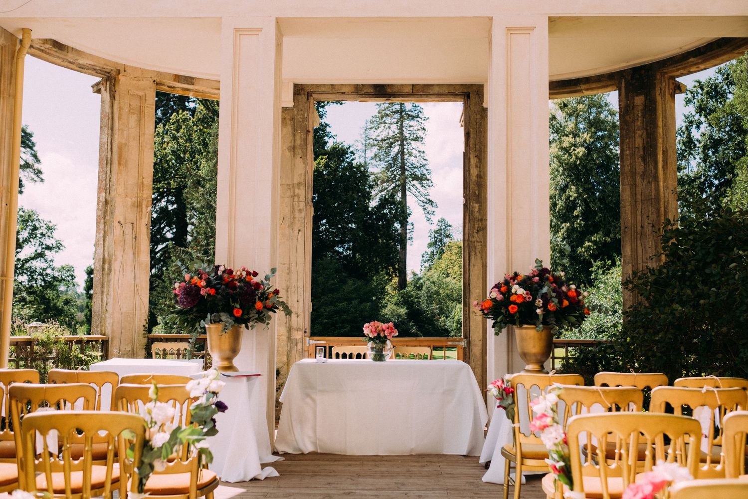An outdoor wedding ceremony setup with wooden chairs and floral arrangements under white pillared columns.
