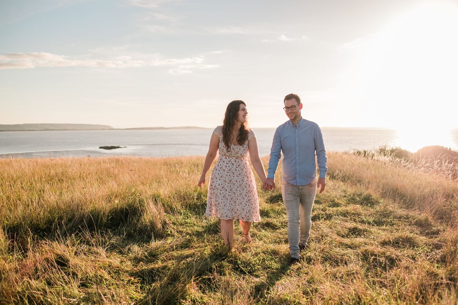 A couple walks through a sunlit meadow during golden hour with ocean views.