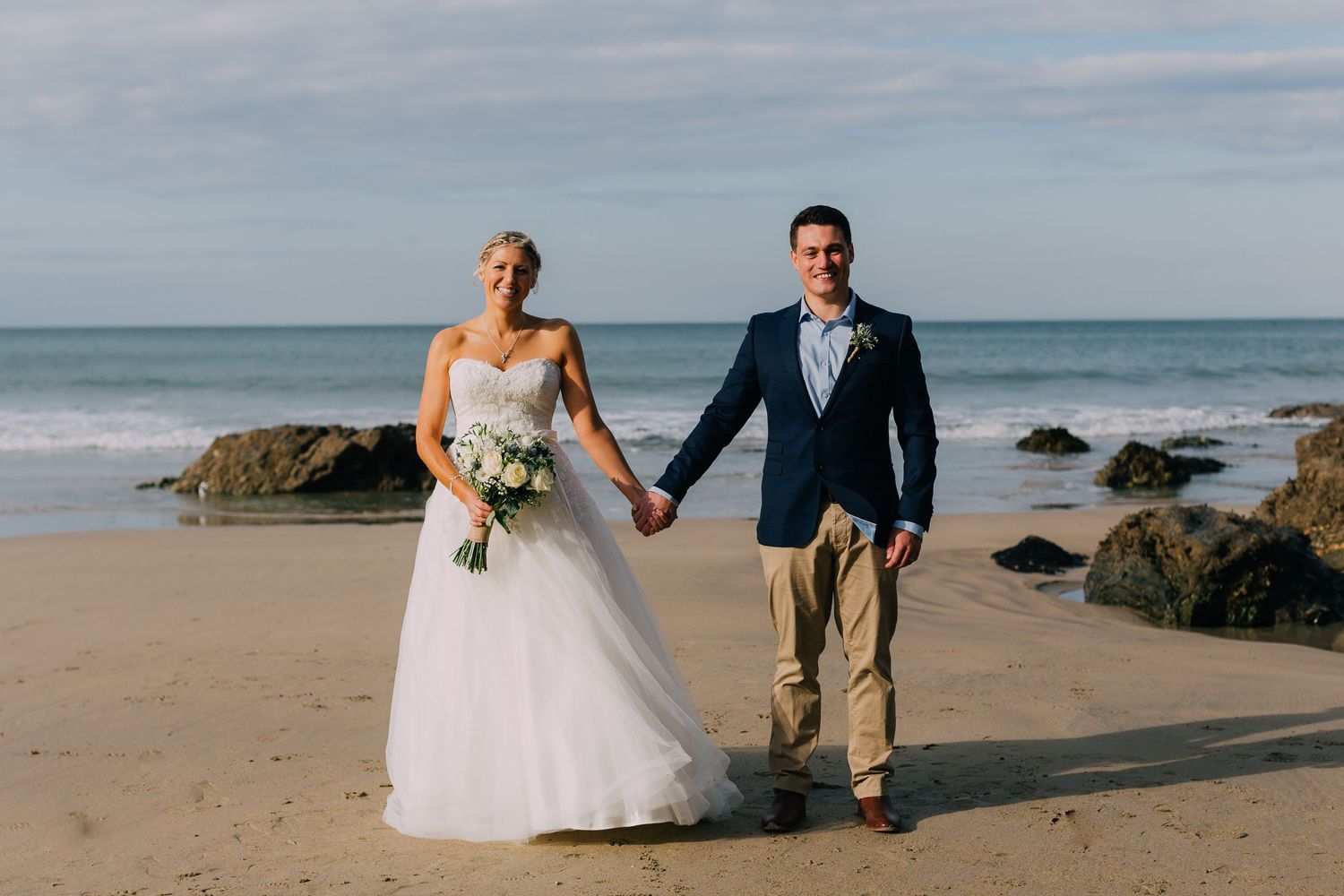A couple in wedding attire walks hand in hand along a scenic beach with rocky outcrops.