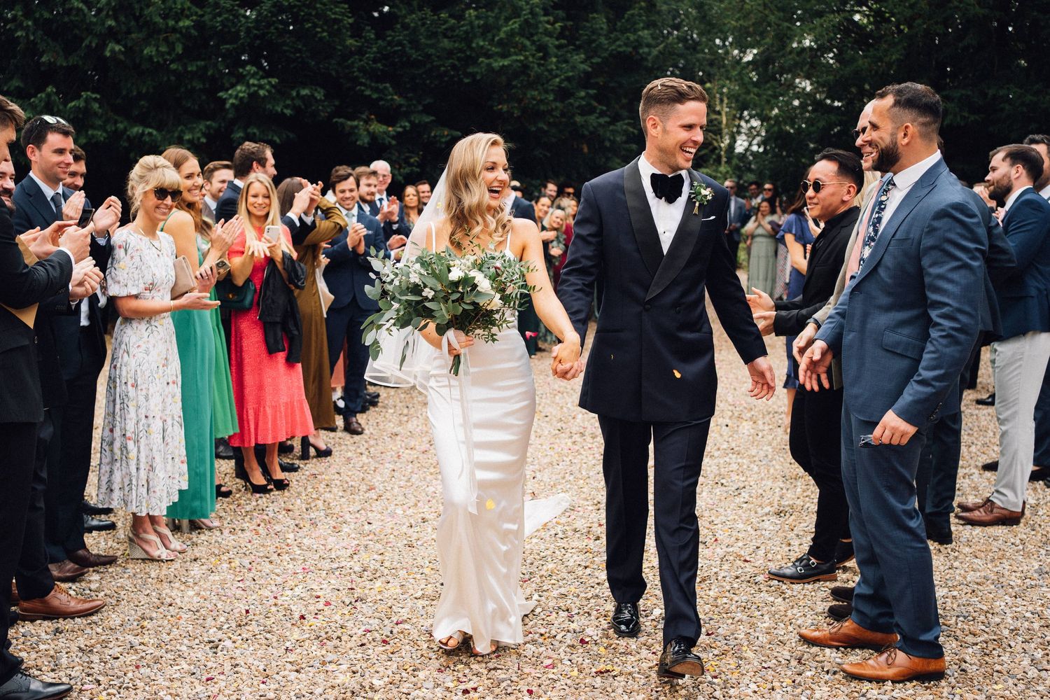 Wedding guests gathered on a gravel path outside surrounded by greenery.