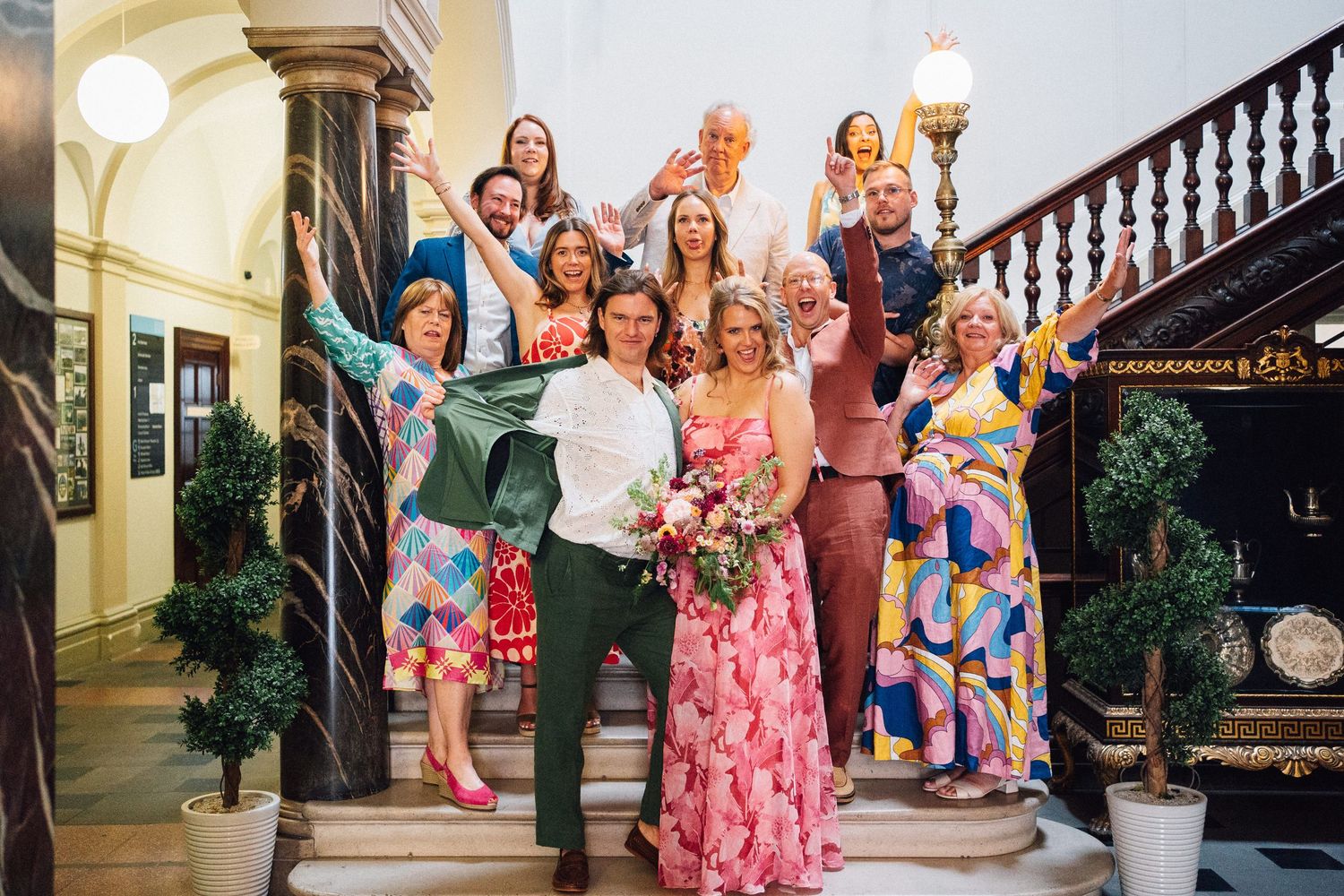 A joyful wedding party poses on grand staircase, wearing colorful outfits, with bride holding pink bouquet.