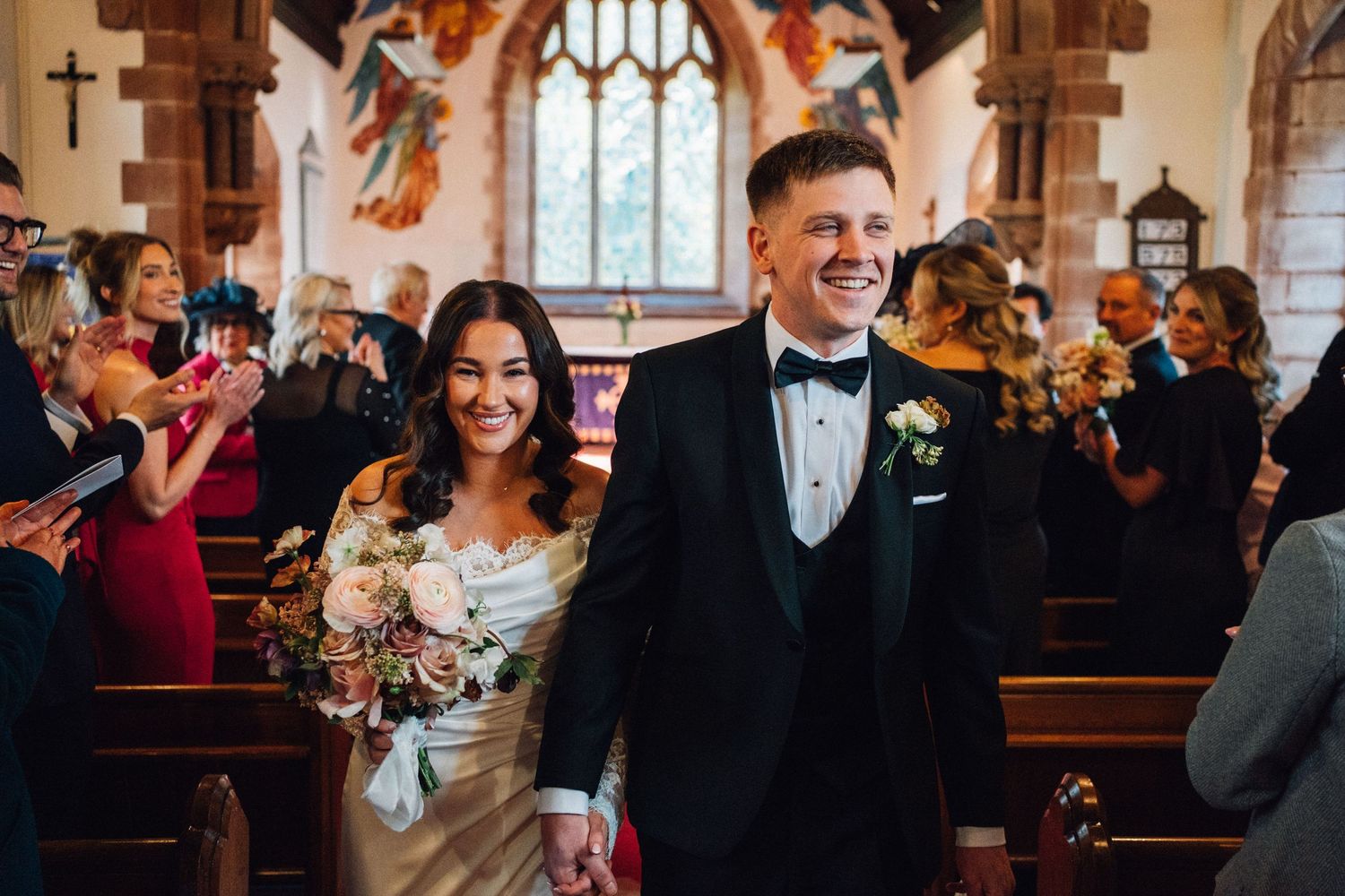 Smiling newlyweds walking hand in hand down the aisle of a decorated church after their ceremony.