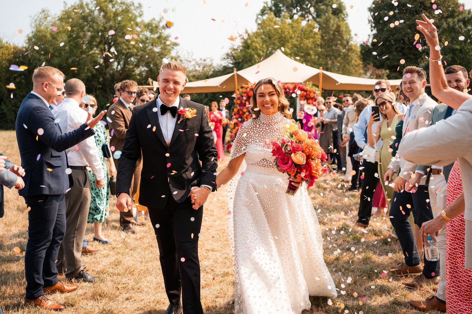 Bride and groom walk through confetti shower surrounded by cheering guests at outdoor wedding celebration.