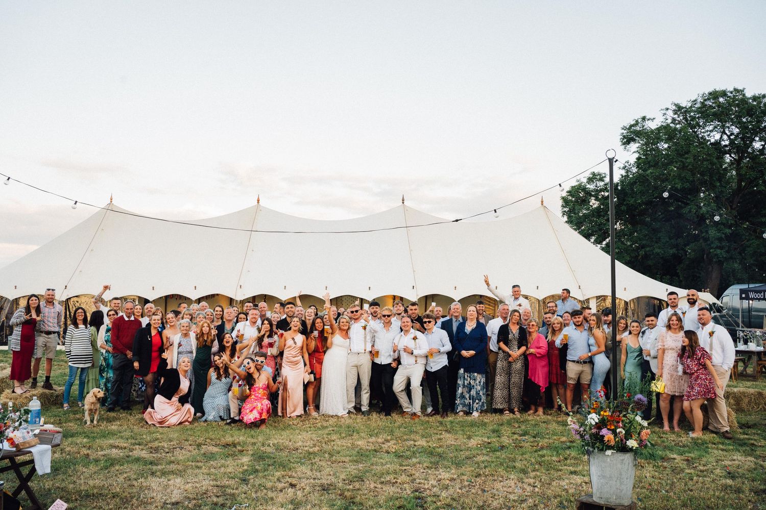 Large group of wedding guests gathered outside a white marquee tent for a panoramic photo in a field.