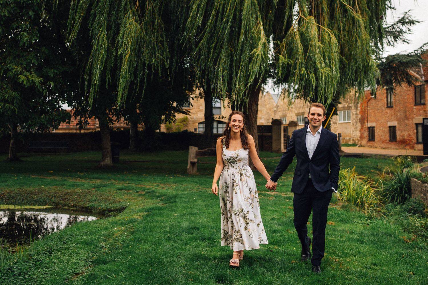 A couple walks together through a lush garden setting with weeping willow trees and historic brick buildings.