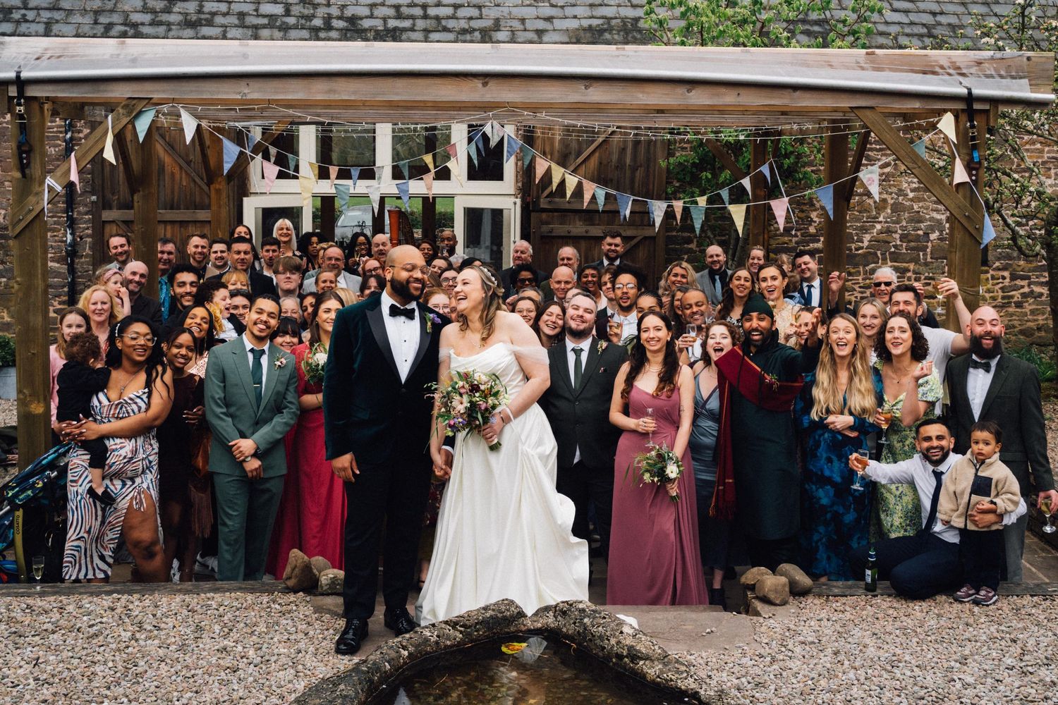 A large group wedding photo of guests and the newlyweds gathered under a bunting-decorated outdoor pavilion.