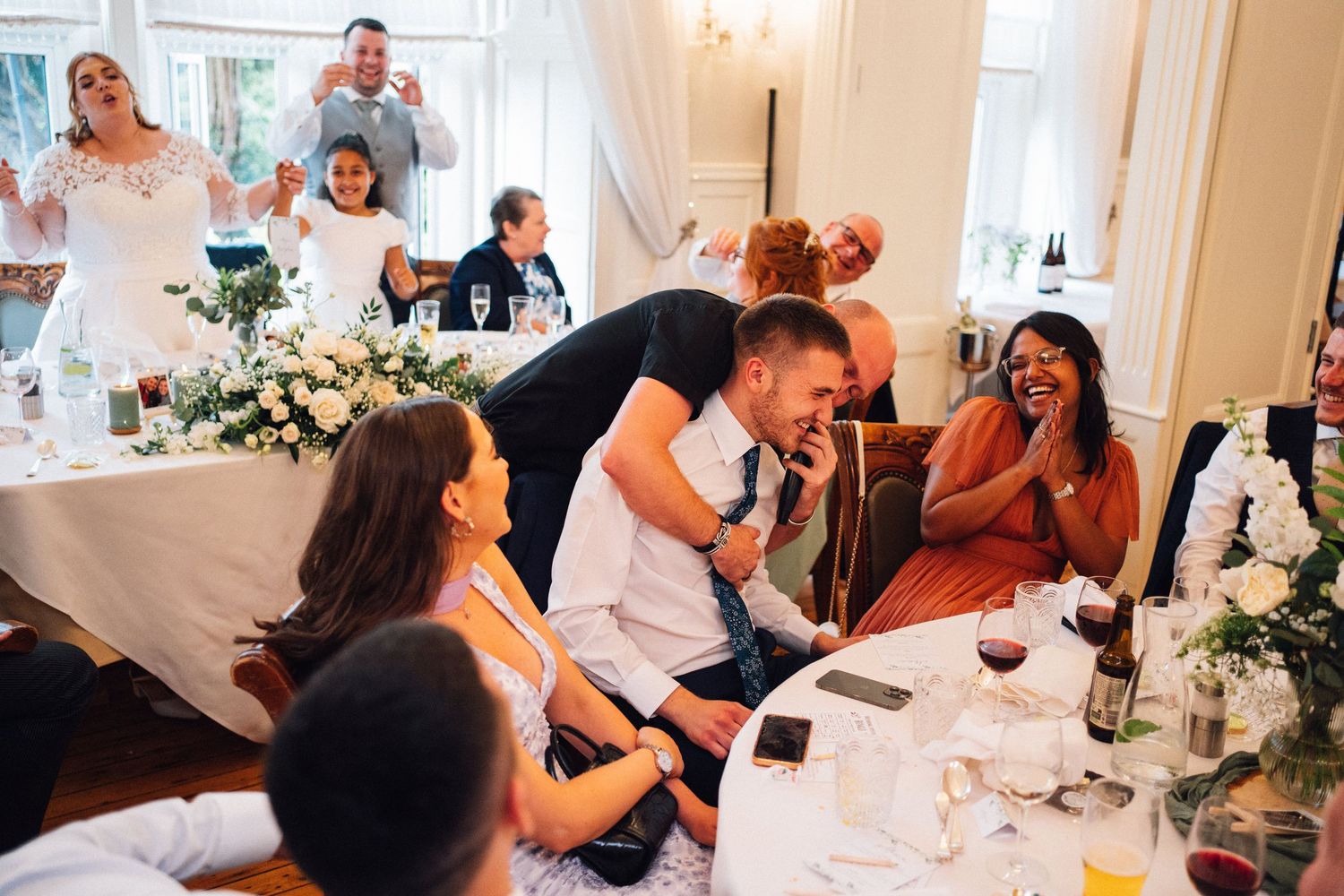 Wedding guests seated at long tables in a bright dining room with white floral centerpieces.