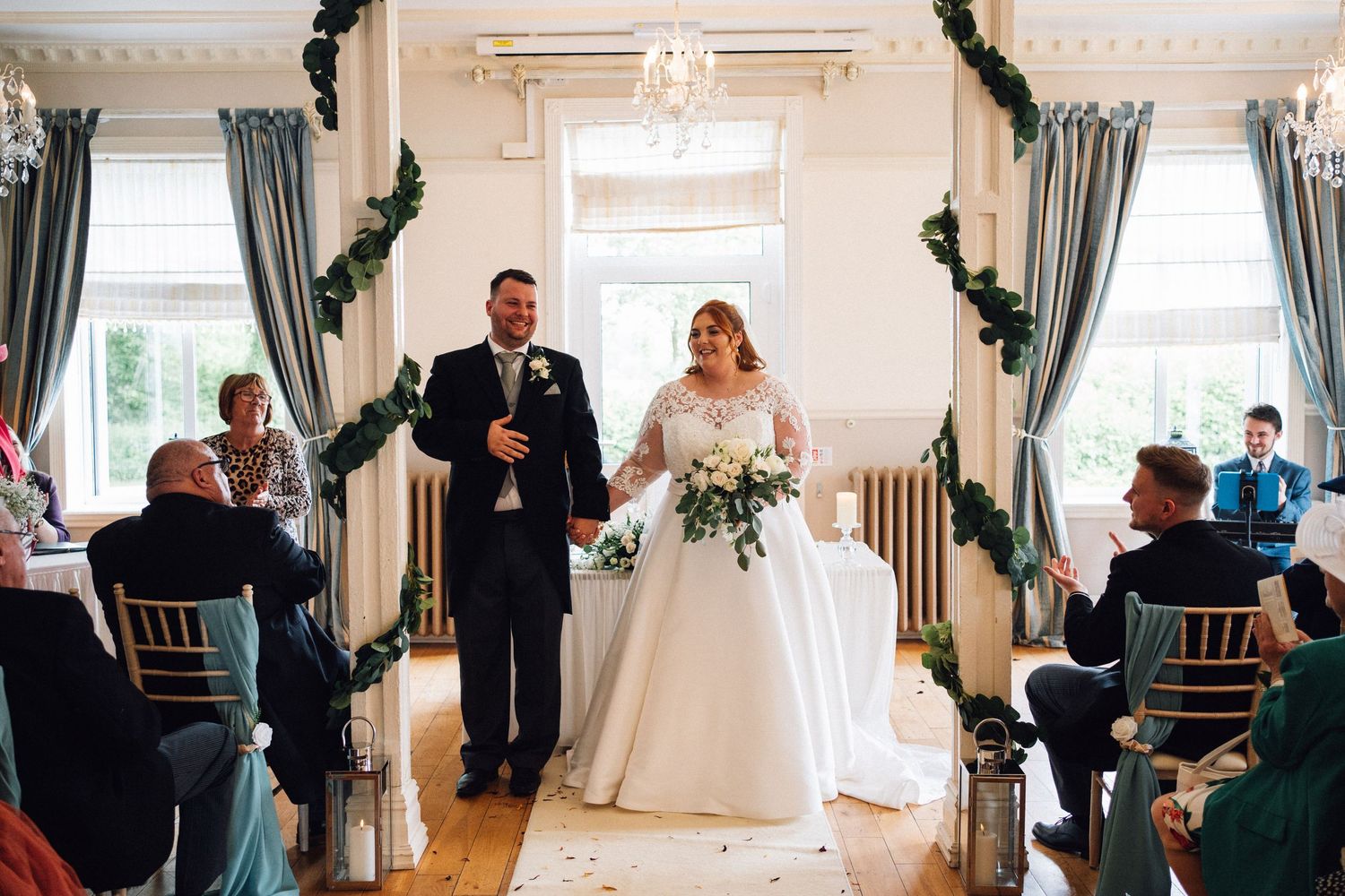 An elegant indoor wedding ceremony takes place in a bright room with chandelier and greenery-draped archway.