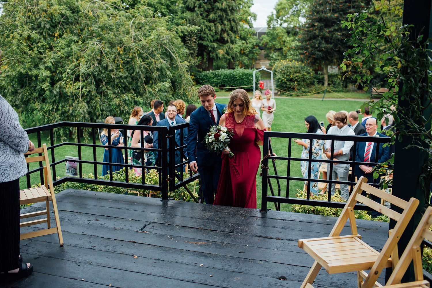 Wedding guests stand on a wooden deck surrounded by lush greenery during an outdoor celebration.