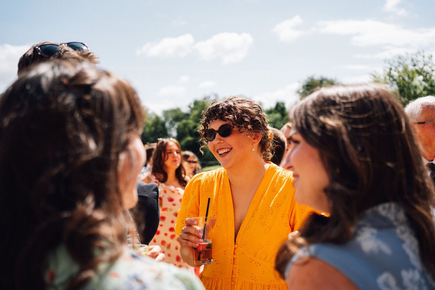 A group of wedding guests chat and laugh together outdoors on a sunny summer day.
