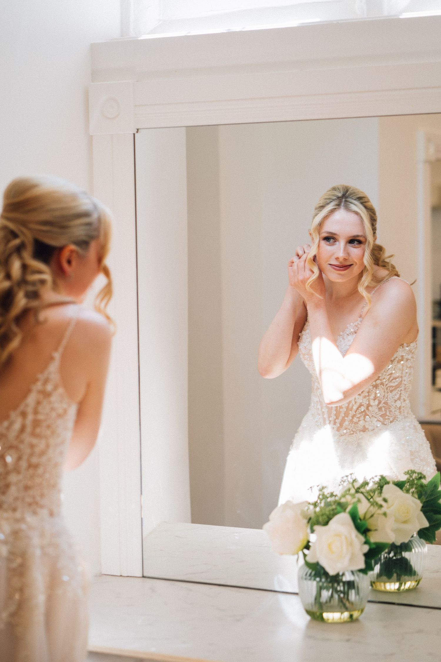 A bride in a lace wedding dress puts on earrings while looking in a mirror with white roses displayed on the counter.