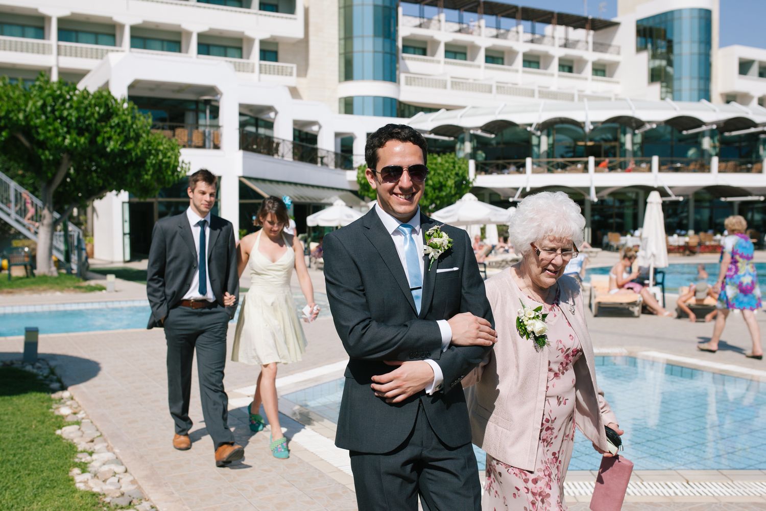 Wedding party members walk and pose near a resort pool area with white buildings.