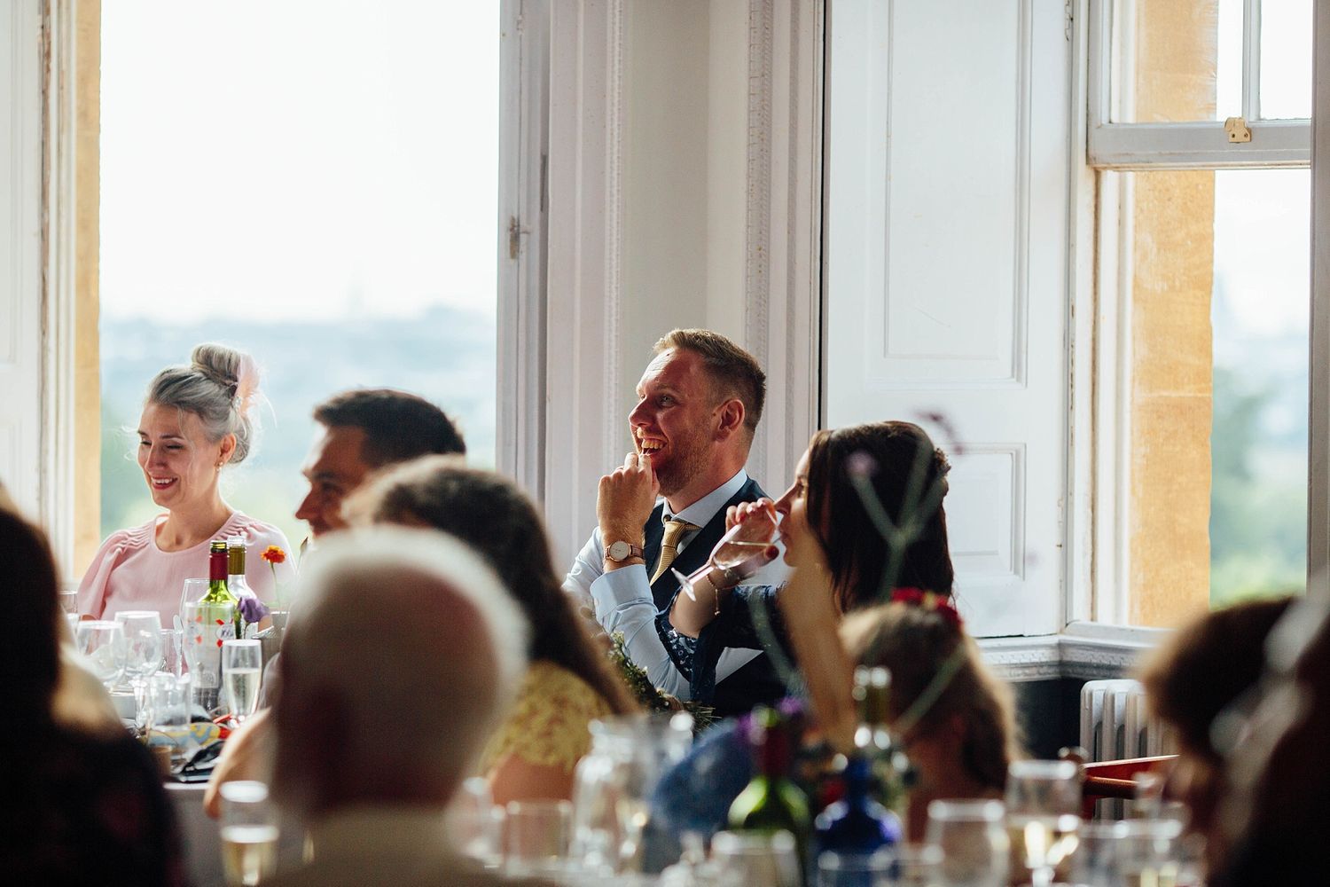 Wedding guests react emotionally during speeches at an elegant reception venue with large windows.