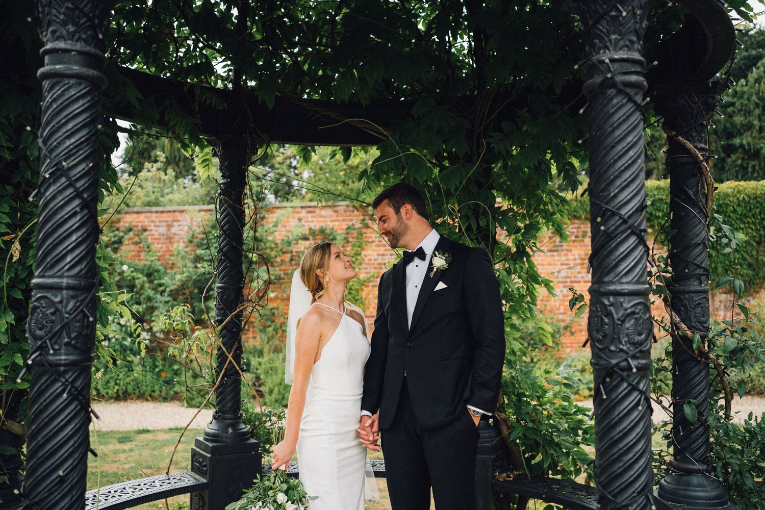Wedding couple stands beneath a vintage iron gazebo with climbing vines and brick wall backdrop.