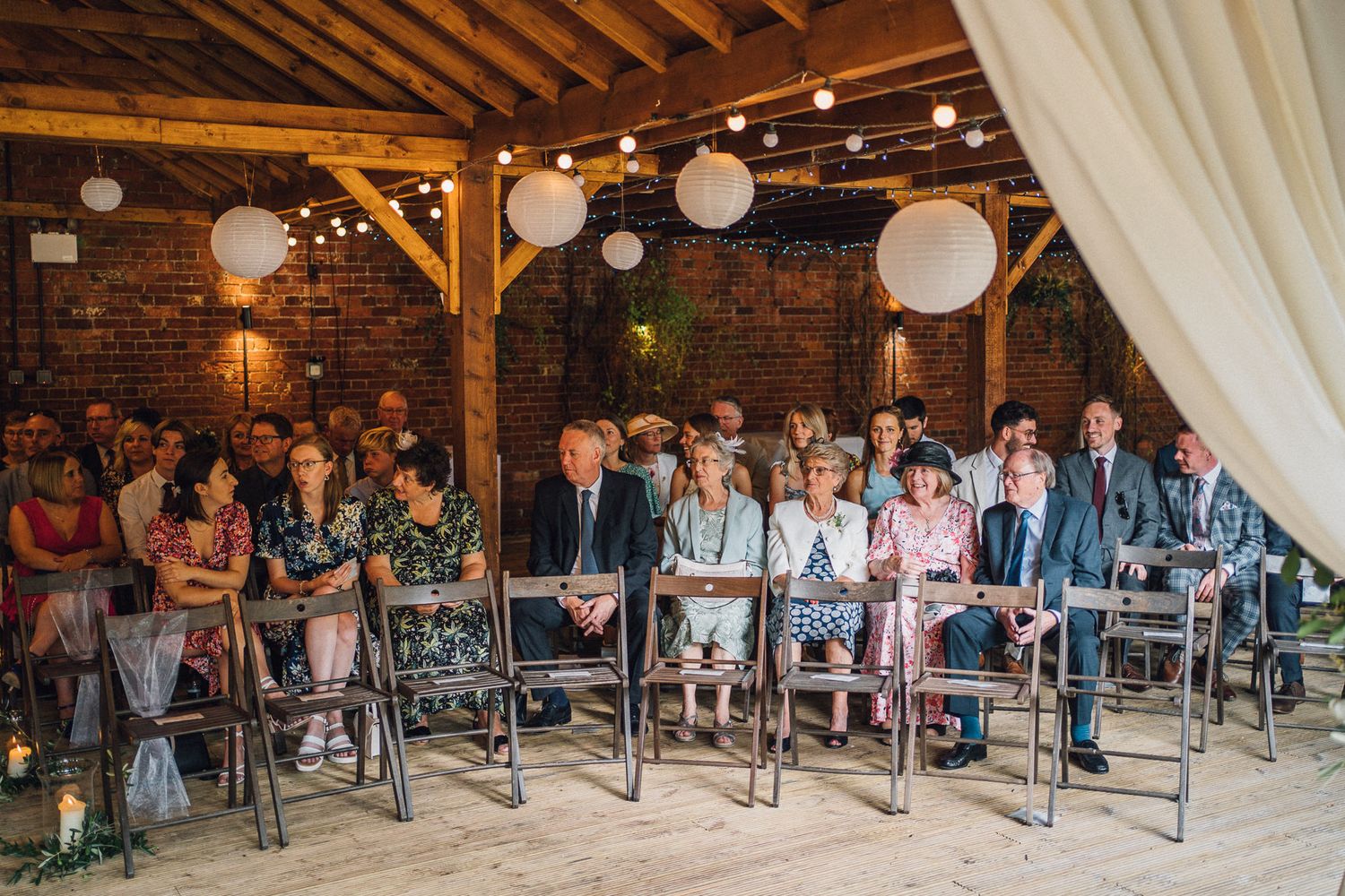 Panoramic view of wedding guests seated in rows inside a rustic barn with string lights and paper lanterns.