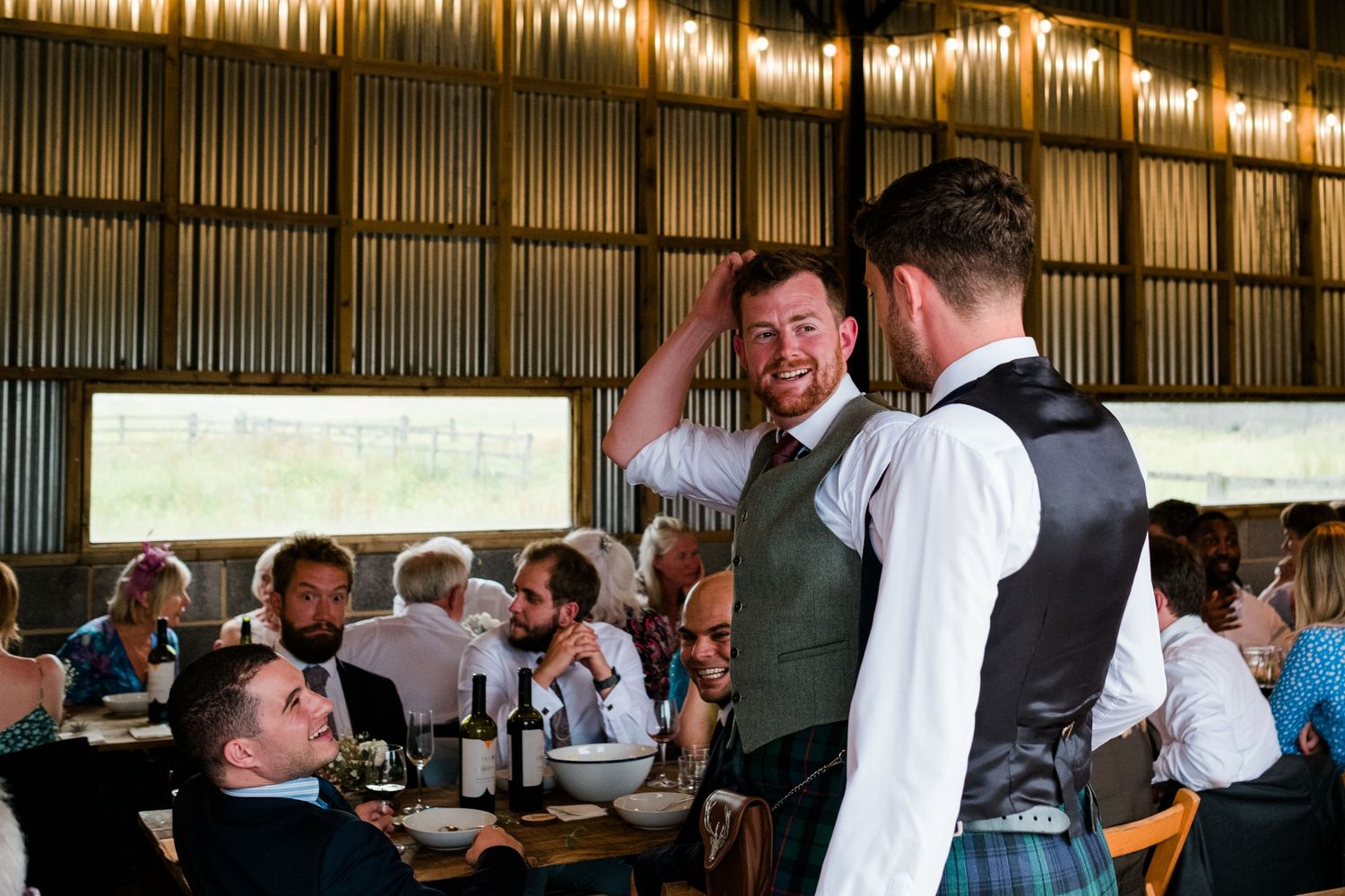 Two people in traditional Scottish attire share a laugh at a wedding reception with guests seated at tables.